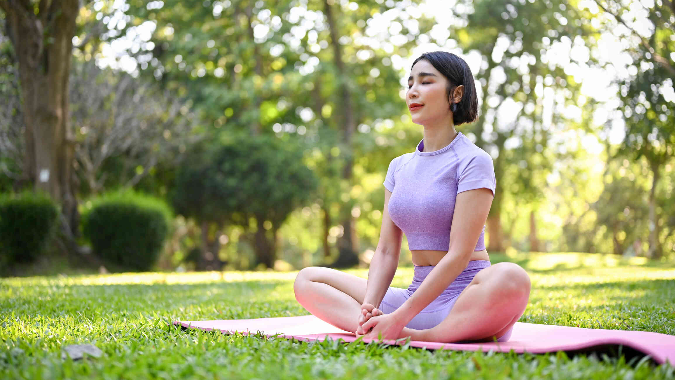 A woman sits cross-legged on a pink yoga mat in a lush green park, meditating with her eyes closed.